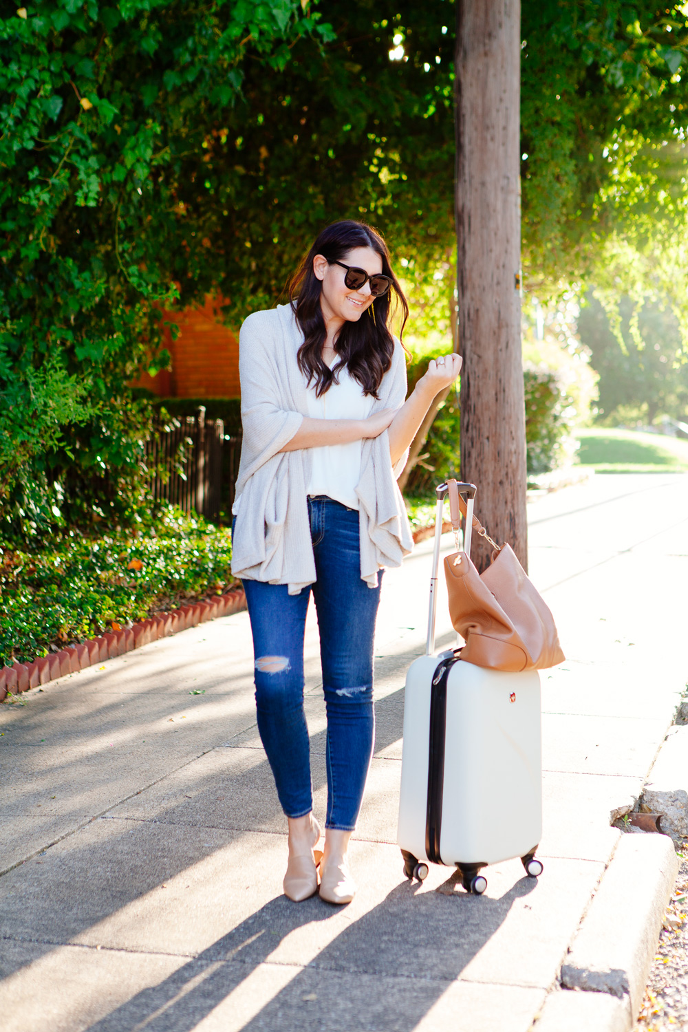 Travel outfit featuring an oatmeal sweater with distressed denim and nude flats, with a white carry-on. As seen on Kendi Everyday